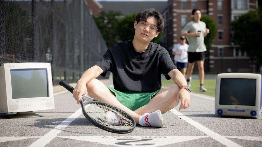 A male student sits on a track between two old computer monitors while holding a tennis racket